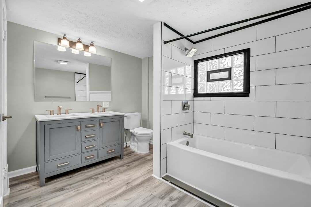 A modern bathroom with a grey double vanity, white subway tile shower, and wood-look flooring — Photo by Josh Lemmon on Unsplash
