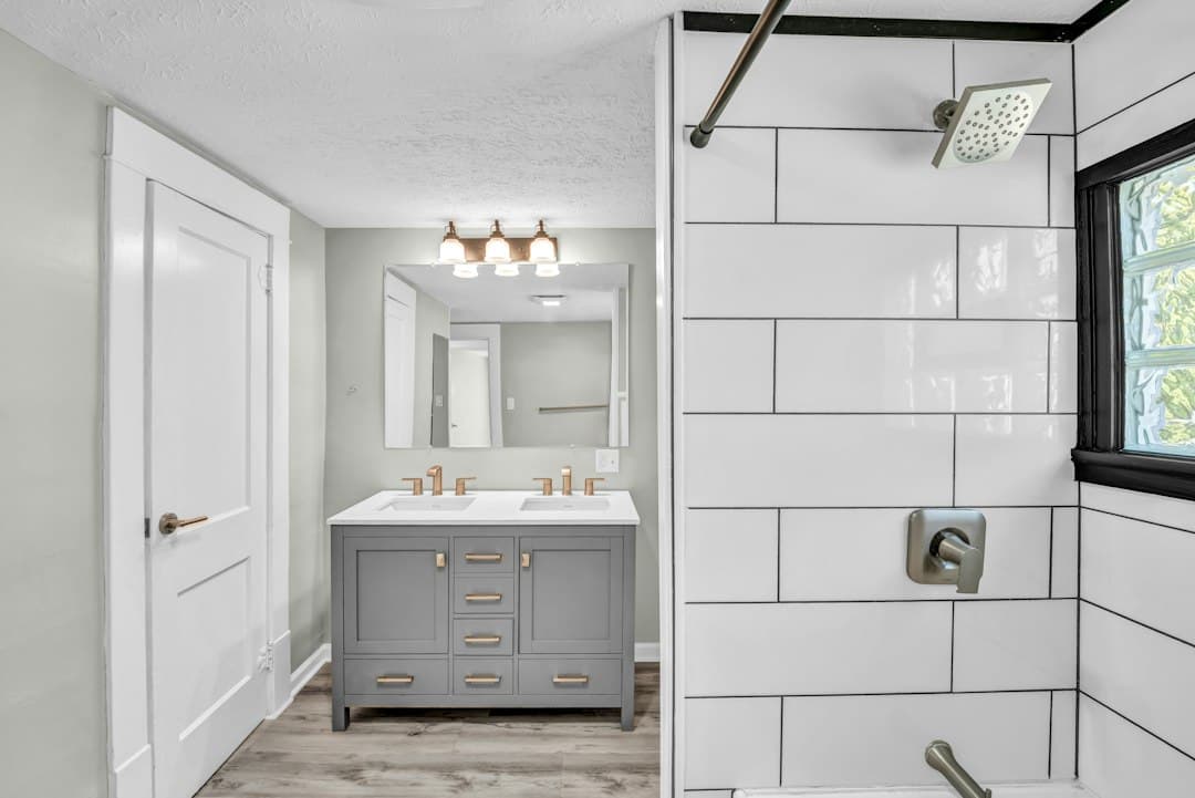 A bathroom with a grey double vanity, white subway tile shower, and light wood flooring — Photo by Josh Lemmon on Unsplash