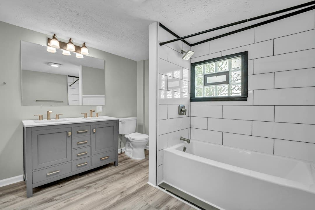 A bathroom with a grey double vanity, a white bathtub, and white subway tile walls — Photo by Josh Lemmon on Unsplash